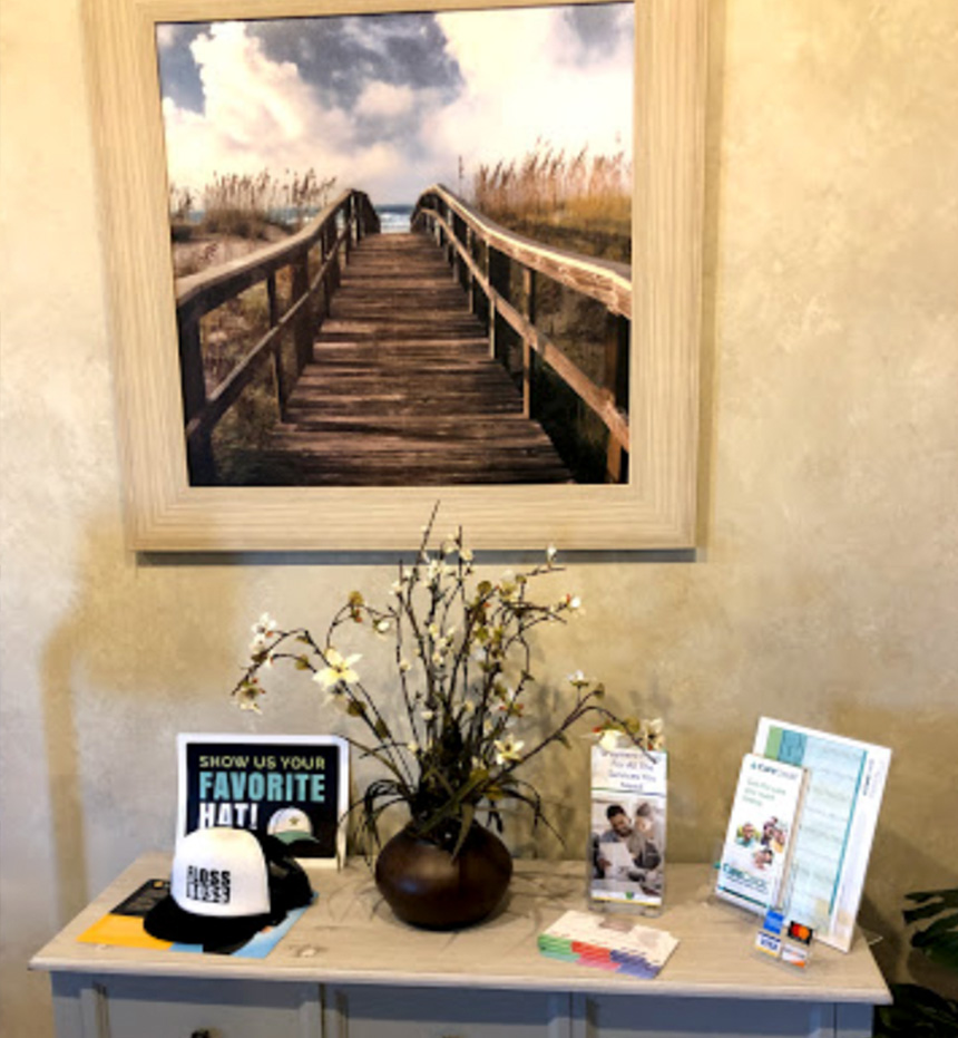 A framed artwork of a coastal scene hangs above a wooden bench, which is adorned with a potted plant and a vase. Below the bench, there s a small table with promotional materials on it.