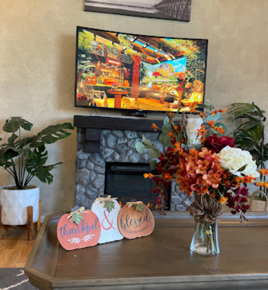 A cozy living room scene with a television displaying a colorful graphic, a fireplace, and decorative items such as pumpkins, flowers, and a sign.