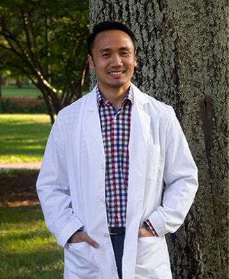 A man in a white lab coat stands confidently outdoors, posing for the camera.