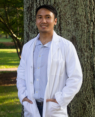 The image shows a man in a white lab coat standing outdoors, smiling at the camera.