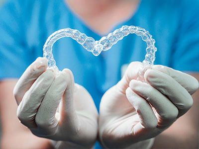 The image shows a person s hands holding a heart-shaped object that resembles a dental retainer or mouthguard, with the background featuring medical gloves and a blue surgical gown.