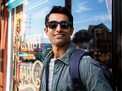 A man in sunglasses is standing outside a store, holding a backpack and smiling.