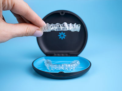 A person s hand is holding a clear plastic tray with a set of white dental implants, which are placed inside a blue and black box labeled  DENTAL IMPLANTS  on a light blue background.