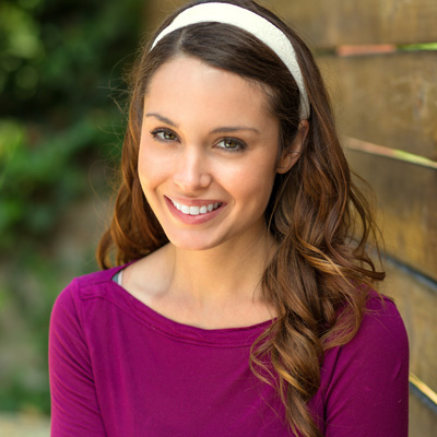Smiling woman with long hair, wearing a purple top and headband, standing in front of a wooden fence.