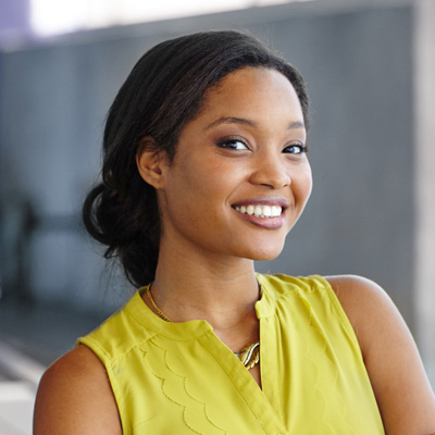 The image features a smiling woman with dark hair, wearing a yellow top and a necklace, posing against a blurred background.