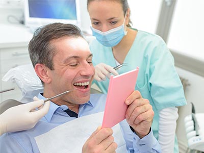 The image is a photograph featuring an adult male seated in a dental chair, holding a tablet with a pink screen and smiling broadly. He appears to be engaged in a conversation or presentation with another person, who is standing behind him and wearing surgical scrubs. The individual standing is also smiling and looking towards the camera. The setting suggests a dental office environment, indicated by the presence of dental equipment and instruments such as a dental mirror and a small model of a mouth. There are no visible texts in the image.