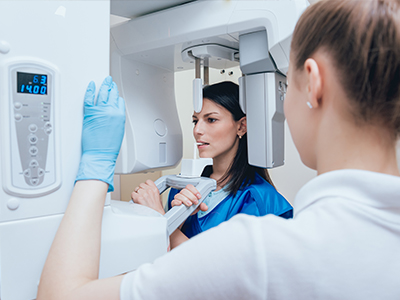 The image features a woman in a blue uniform standing next to a large, modern-looking 3D scanner, with another person wearing gloves and a face mask interacting with the machine.