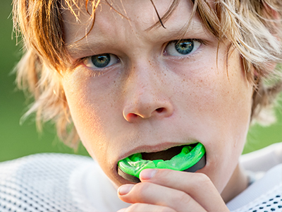 The image shows a young male with blonde hair, wearing a sports jersey, holding a green object near his mouth.