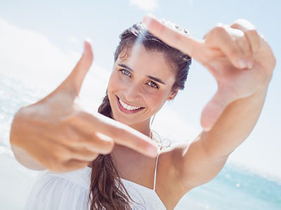 A young woman with a radiant smile is seen holding up her index finger against the bright backdrop of the ocean under a clear sky.